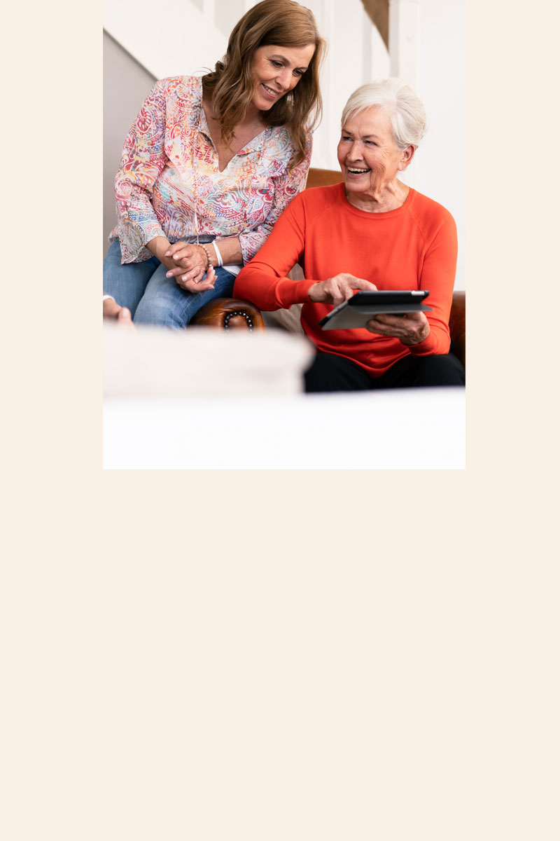 Ältere Frau zeigt lächelnd etwas auf einem Tablet, während eine jüngere Frau daneben steht und hilft