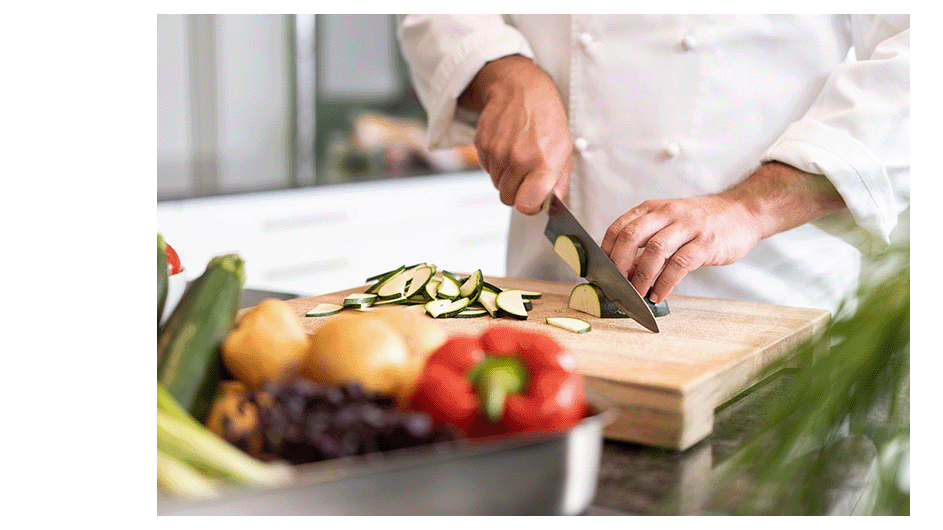 Koch in weißer Jacke schneidet Gemüse auf einem Holzbrett