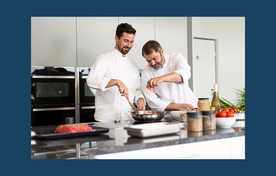 Zwei Köche bereiten  in einer Küche ein Pfannengericht  zu. Auf der Arbeitsplatte stehen frische Zutaten und Gewürze bereit
