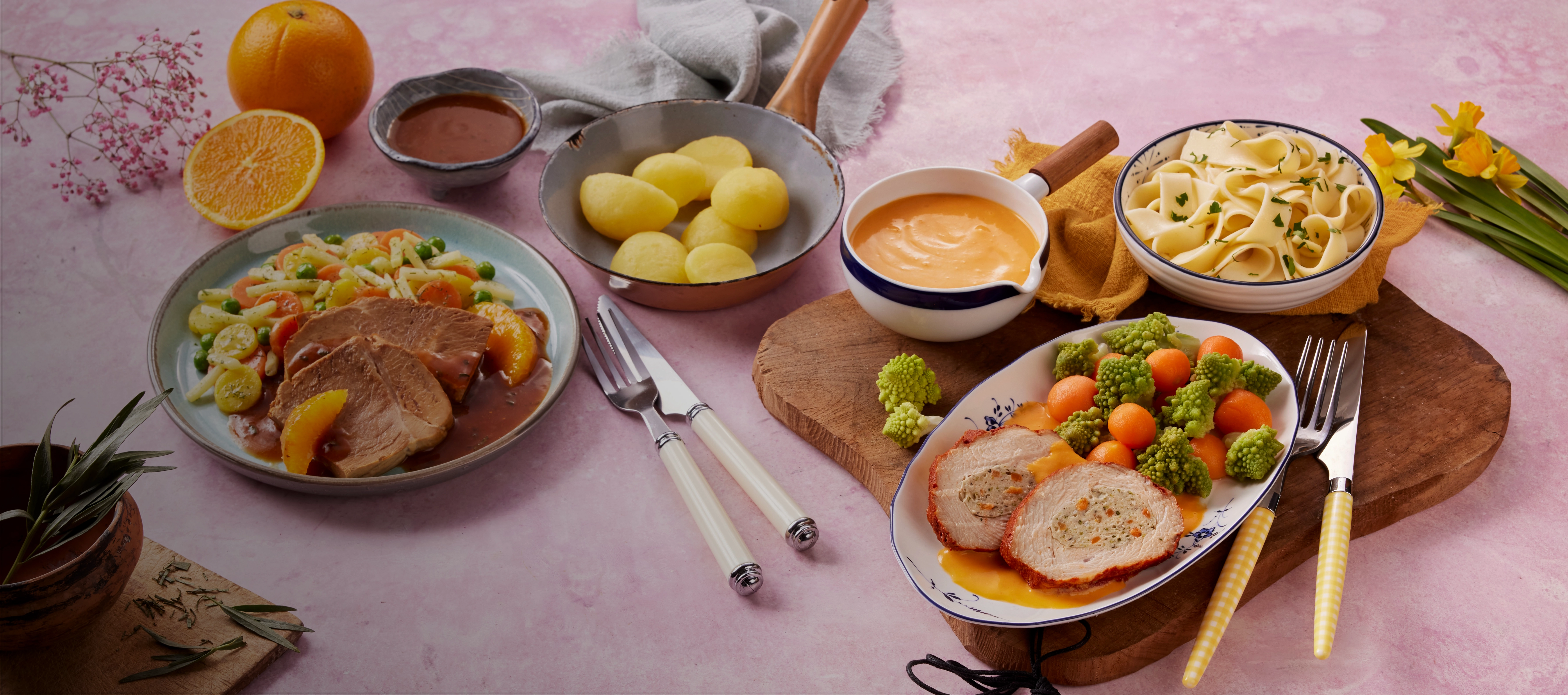 Frühlingshaft gedeckter Tisch mit zwei Tellern leichter Fleischgerichte, frischem Gemüse, Kartoffeln, Nudeln und Soßen, dekoriert mit verschiedenen Blumen