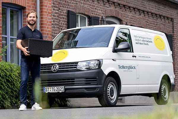 lachender Mann steht mit Isolierbox neben einem Tellerglück-Lieferwagen