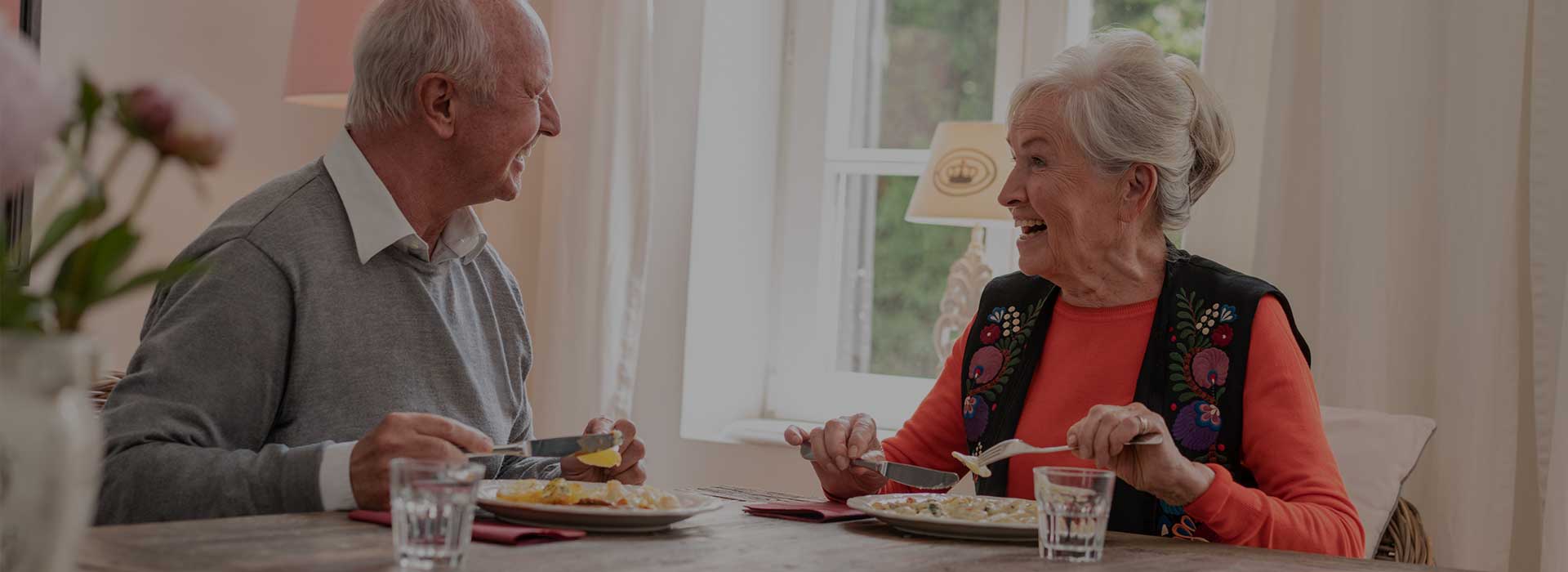 Ein älteres Ehepaar sitzt gemeinsam beim Essen am Tisch und lacht sich gegenseitig an