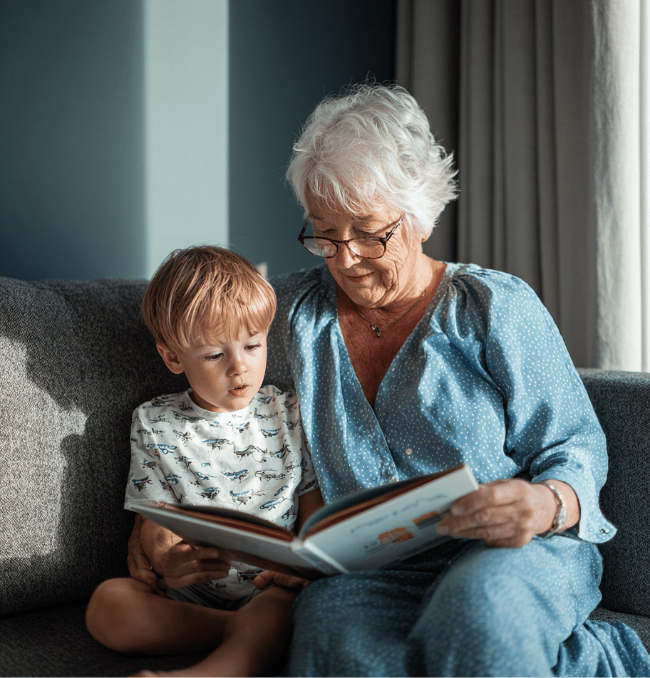 Eine Oma sitzt mit ihrem Enkel in einem grauen Stuhl. Sie liest dem kleinen Kind etwas aus einem Buch vor. 