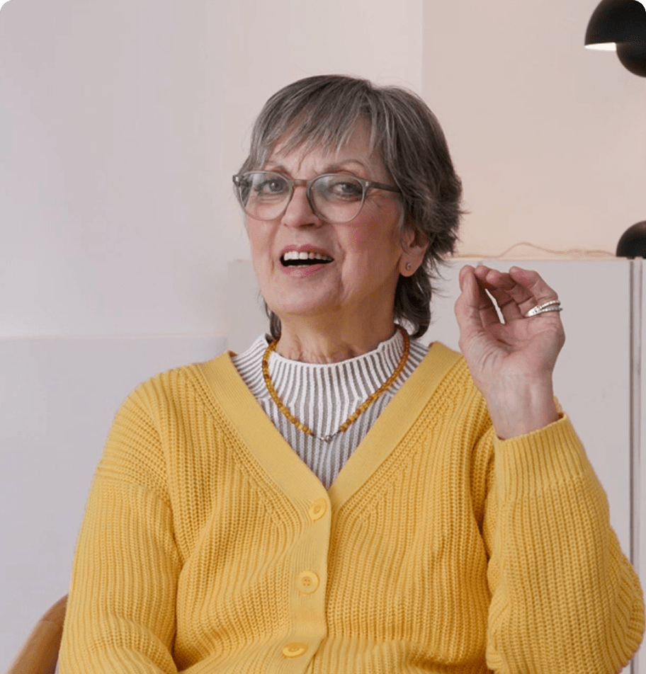 Person in einer gelben Strickjacke hebt leicht eine Hand, im Hintergrund moderne Raumbeleuchtung.