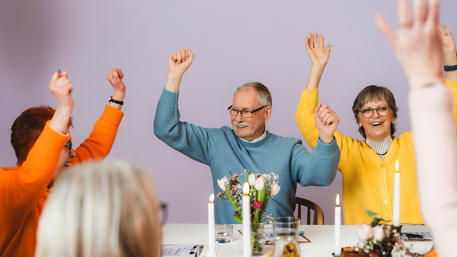Seniorengruppe sitzt gemeinsam am Tisch und hebt fröhlich die Arme in die Höhe.