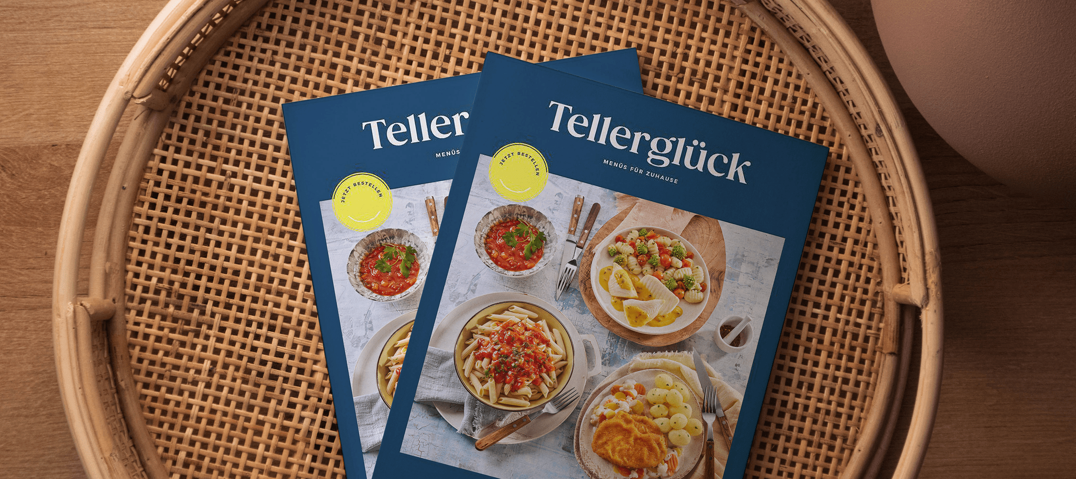Auf einem geflochtenem Tablet liegen zwei Kataloge des neuen Tellerglück-Sortiments.