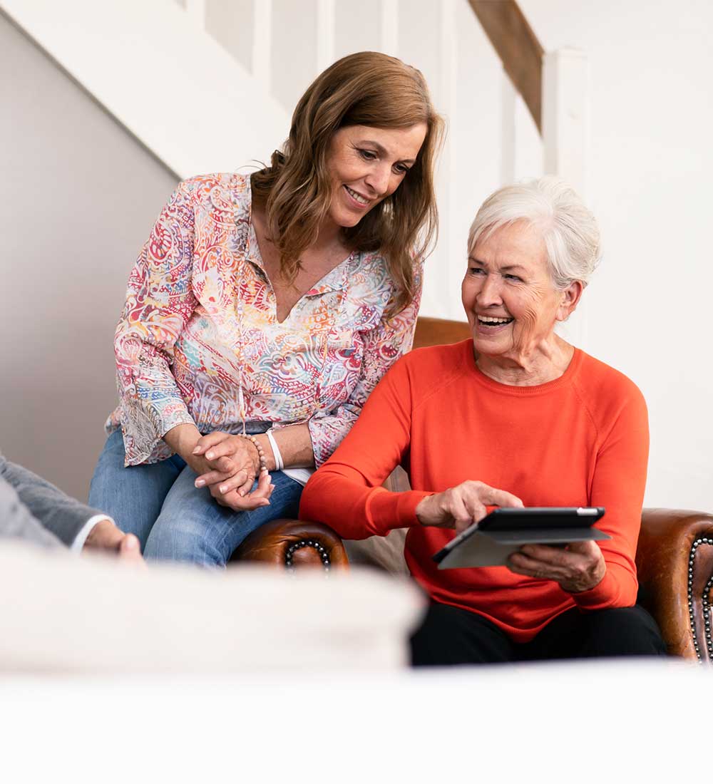 Ältere Frau zeigt lächelnd etwas auf einem Tablet, während eine jüngere Frau daneben steht und hilft
