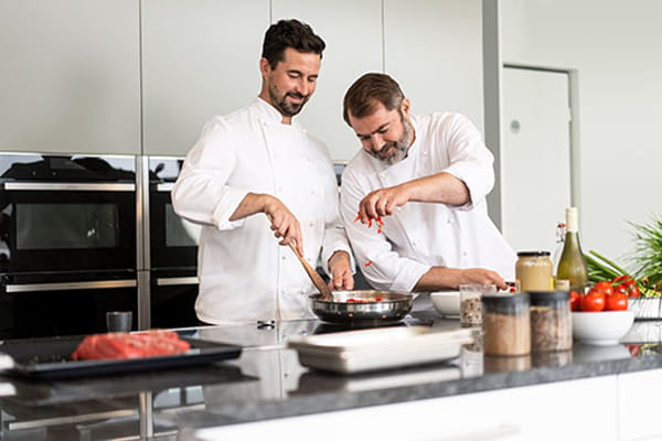 Zwei Köche bereiten in einer Küche ein Pfannengericht zu. Auf der Arbeitsplatte stehen frische Zutaten und Gewürze bereit