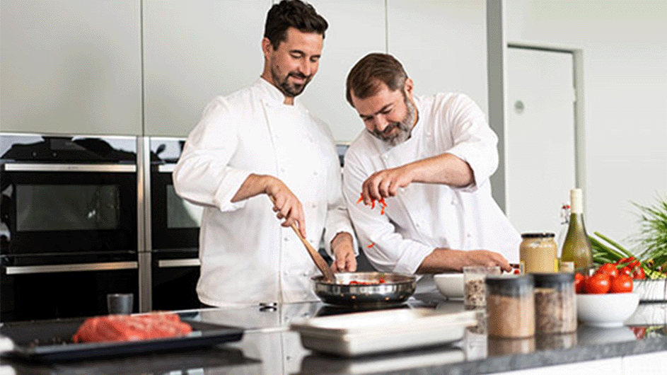 Zwei Köche bereiten  in einer Küche ein Pfannengericht  zu. Auf der Arbeitsplatte stehen frische Zutaten und Gewürze bereit