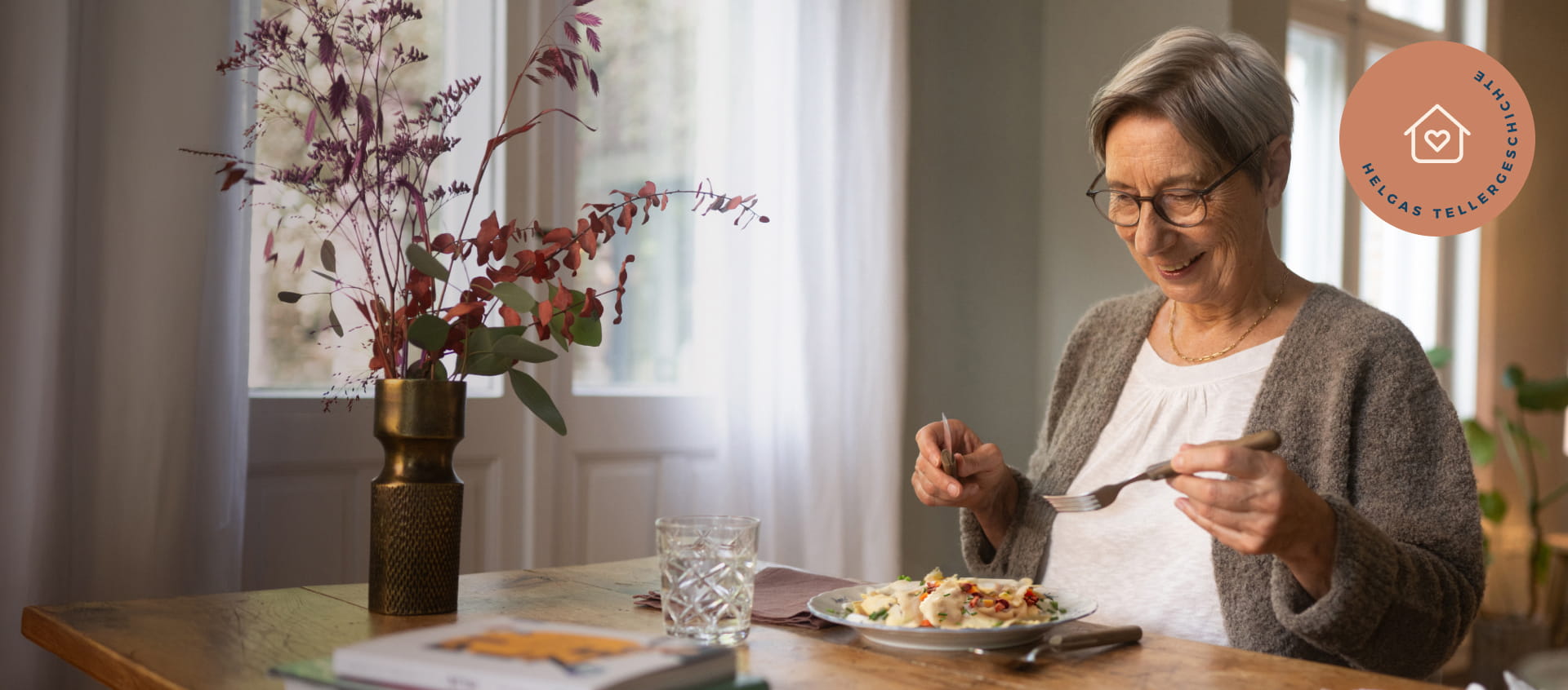 Ältere Dame sitzt an einem gedeckten Tisch. Sie hält Besteck in der Hand und schaut glücklich auf ihren gefüllten Teller.