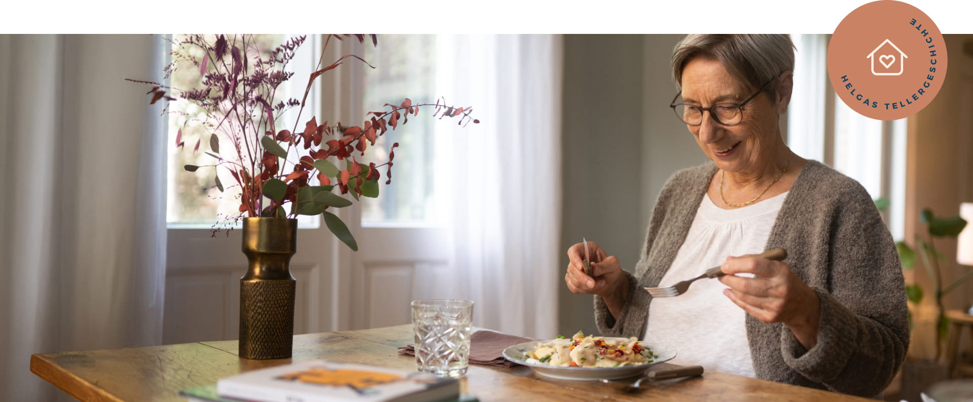 Ältere Dame sitzt an einem gedeckten Tisch. Sie hält Besteck in der Hand und schaut glücklich auf ihren gefüllten Teller.