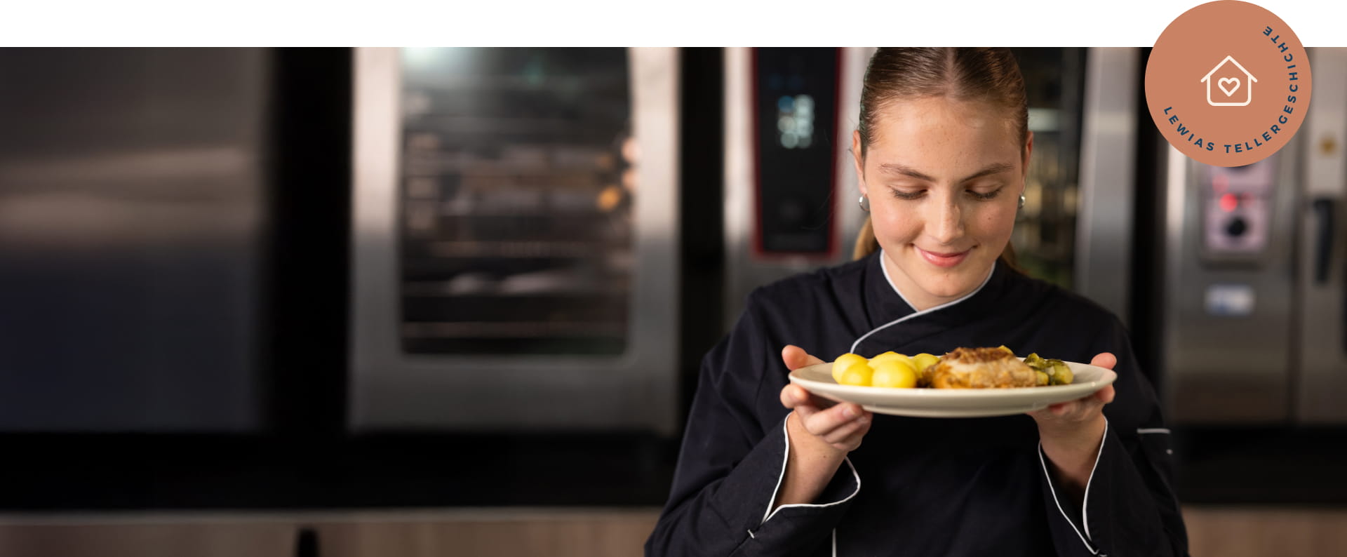 Junge Köchin hält einen Teller mit einem Menü in beiden Händen. Sie hält den Teller etwas erhöht und schaut auf das Gericht. Sie steht in einer Küche.