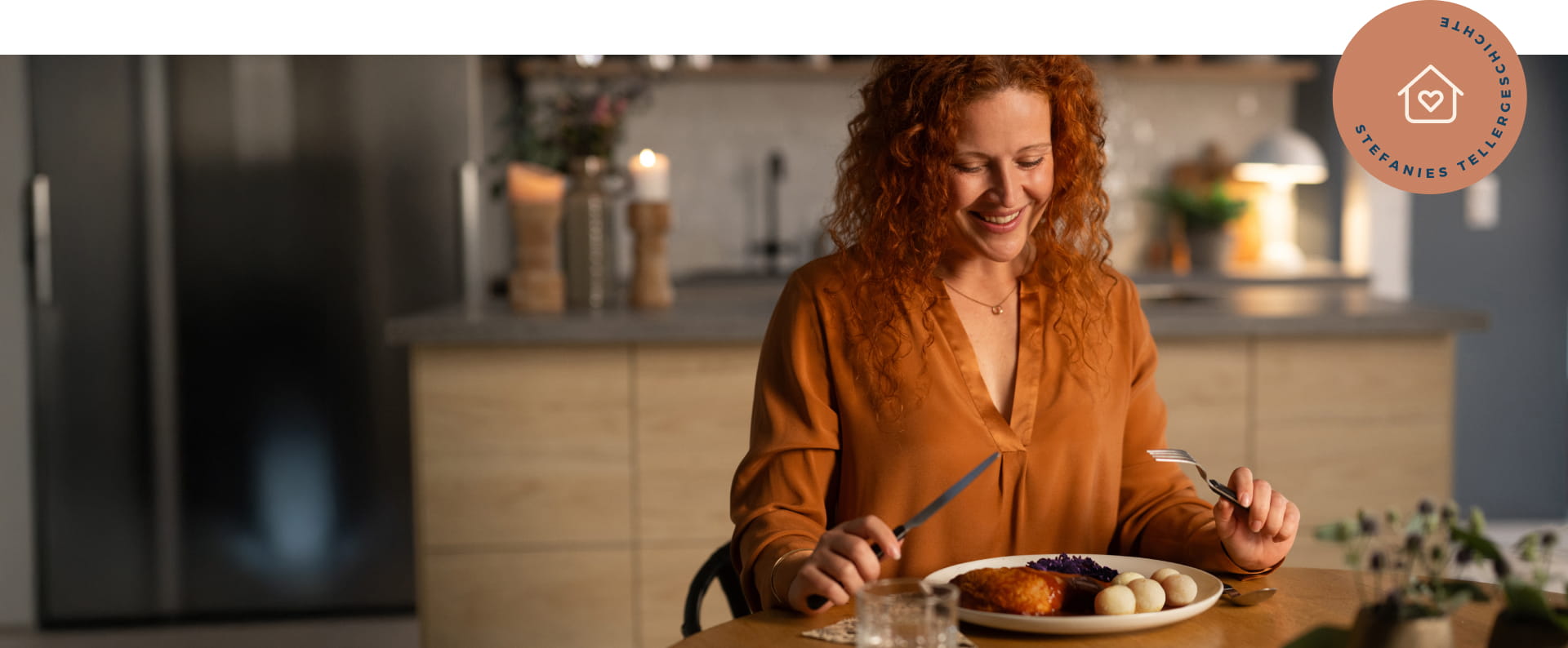 Jüngere Frau sitzt an einem Wohzimmertisch und hält Besteck in der Hand. Sie blickt auf den Teller, der vor ihr auf dem Holztisch steht und lächelt.