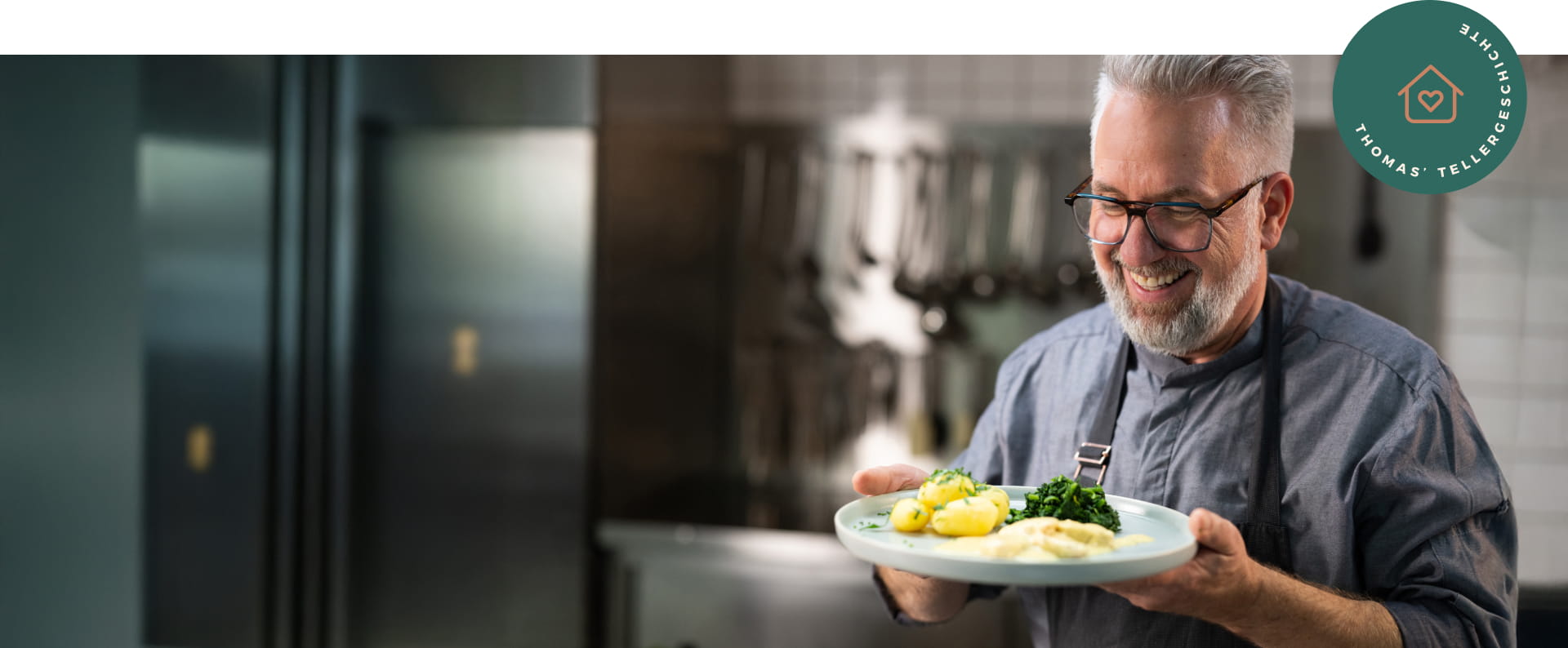 Männlicher Koch steht in einer Industrieküche, lacht und hält einen Teller mit einem Gericht in beiden Händen.