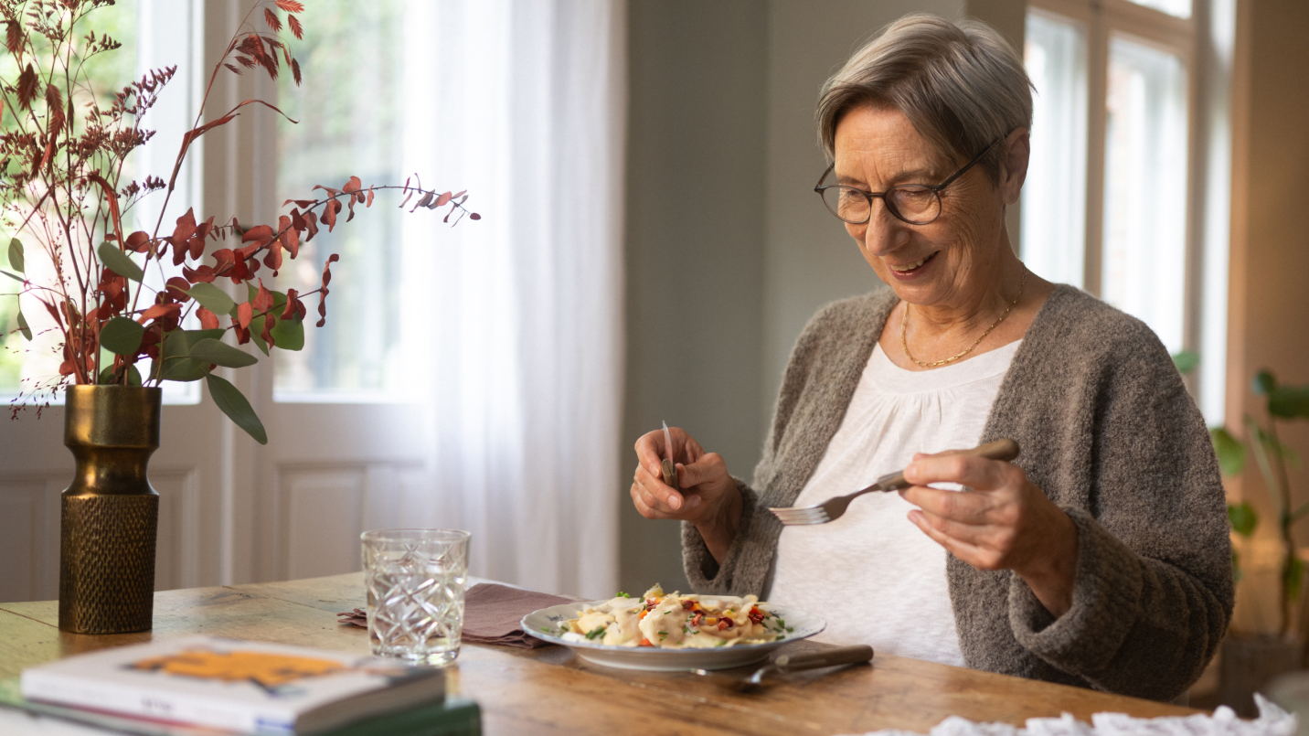 Ältere Frau mit grauen Haaren, Brille und grauer Strickjacke sitzt lächelnd am Holztisch, isst mit Gabel und Messer Ravioloni in cremiger Soße mit Gemüse. Vor ihr Glas Wasser, Buchstapel und Vase mit herbstlichen Blumen. Im Hintergrund helles Fenster mit Vorhang.