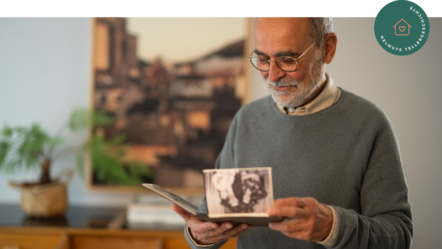 Älterer Mann mit Brille, Bart und grauem Pullover sitzt nachdenklich am Tisch und betrachtet ein aufgeschlagenes Fotoalbum mit Schwarz-Weiß-Fotos. Im Hintergrund grüne Pflanze in Korb und Gemälde einer Stadt. Rechts oben rundes grünes Badge mit Icon „Haus mit Herz“ und Text „HELMUTS TELLERGESCHICHTE“.