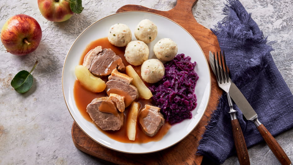 Weißer Teller auf Holzbrett mit geschnittener Gänsebrust in Apfel-Preiselbeersoße, Rotkohl, Speckklößen und Apfelstücken. Links zwei Äpfel, rechts Besteck auf blauem Tuch.