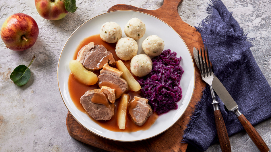 Weißer Teller auf Holzbrett mit geschnittener Gänsebrust in Apfel-Preiselbeersoße, Rotkohl, Speckklößen und Apfelstücken. Links zwei Äpfel, rechts Besteck auf blauem Tuch.