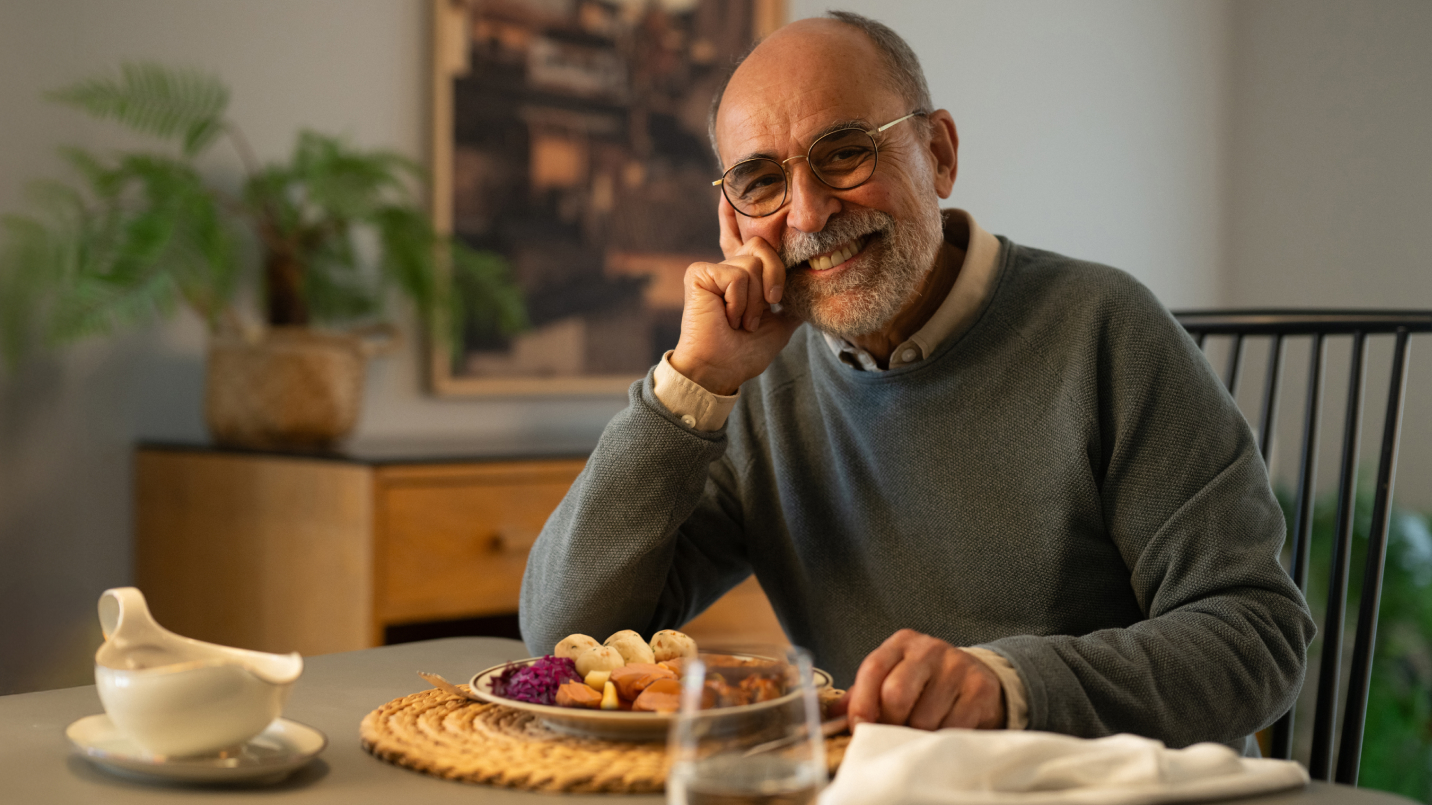 Älterer Mann mit grauem Bart, Brille und grauem Pullover sitzt lächelnd am Tisch, stützt Kinn auf Hand. Vor ihm Teller mit Gänsebrust, Rotkohl, Klößen und Apfelstücken in Soße, daneben Soßenkännchen und Glas. Im Hintergrund Holzschrank, Pflanze und Gemälde einer Stadt.