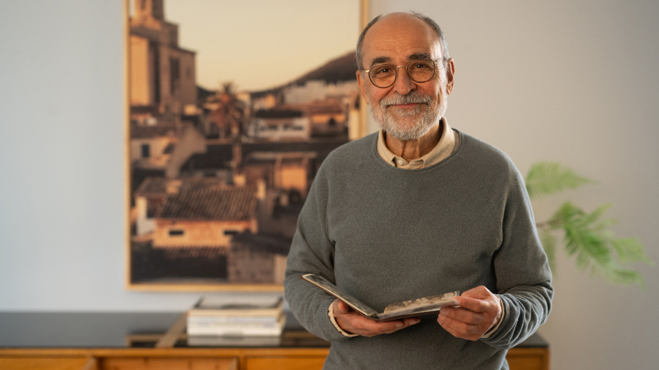Älterer Mann mit grauem Bart, Brille und grauem Pullover steht lächelnd in einem hellen Raum, hält ein aufgeschlagenes Fotoalbum in den Händen. Im Hintergrund gerahmtes Bild einer südländischen Stadt mit Häusern und Kirchturm, links Stapel Bücher auf Holzschrank, rechts grüne Pflanze.