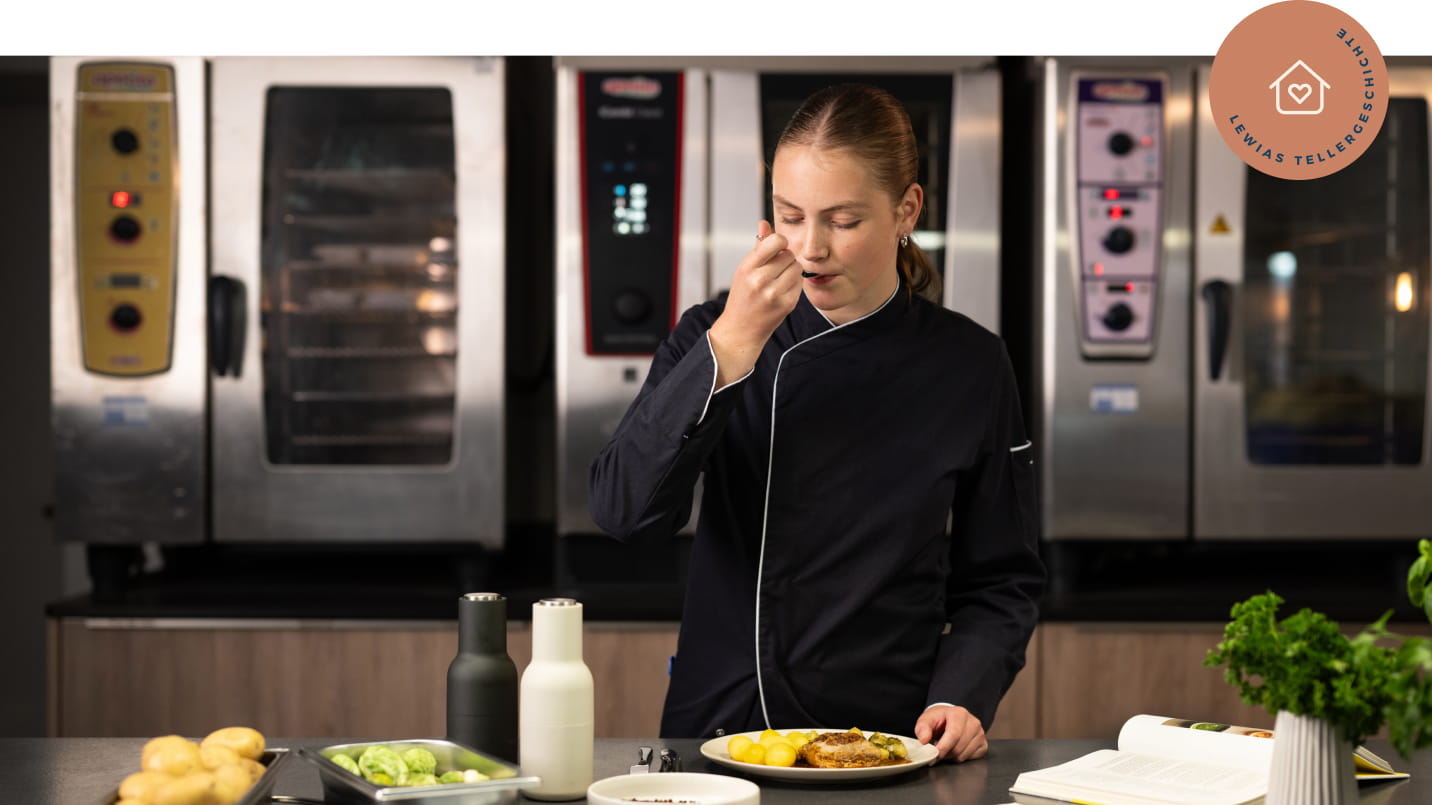 Junge Köchin in schwarzer Uniform mit weißem Kragen steht in Profiküche, hält Teller mit Kartoffeln, paniertem Fleisch und Gemüse, kostet mit der anderen Hand. Vor ihr Gewürzmühlen, Rosenkohl, Brot und offenes Rezeptbuch. Im Hintergrund Edelstahlöfen. Rechts oben roséfarbenes rundes Badge mit Icon „Haus mit Herz“ und Text „LEWIAS TELLERGESCHICHTE“.