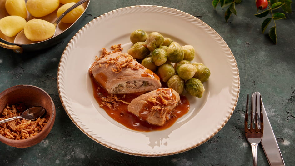 Weißer Teller mit Schweineroulade in Bratenjus, garniert mit Röstzwiebeln, dazu Rosenkohl und Salzkartoffeln. Links Schale mit weiteren Kartoffeln, kleine Schüssel mit Röstzwiebeln und Löffel; rechts Besteck. Hintergrund grünlicher Tisch mit Hagebuttenzweig.