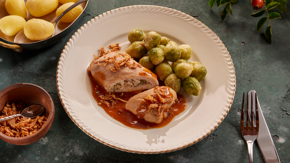 Weißer Teller mit Schweineroulade in Bratenjus, garniert mit Röstzwiebeln, dazu Rosenkohl und Salzkartoffeln. Links Schale mit weiteren Kartoffeln, kleine Schüssel mit Röstzwiebeln und Löffel; rechts Besteck. Hintergrund grünlicher Tisch mit Hagebuttenzweig.