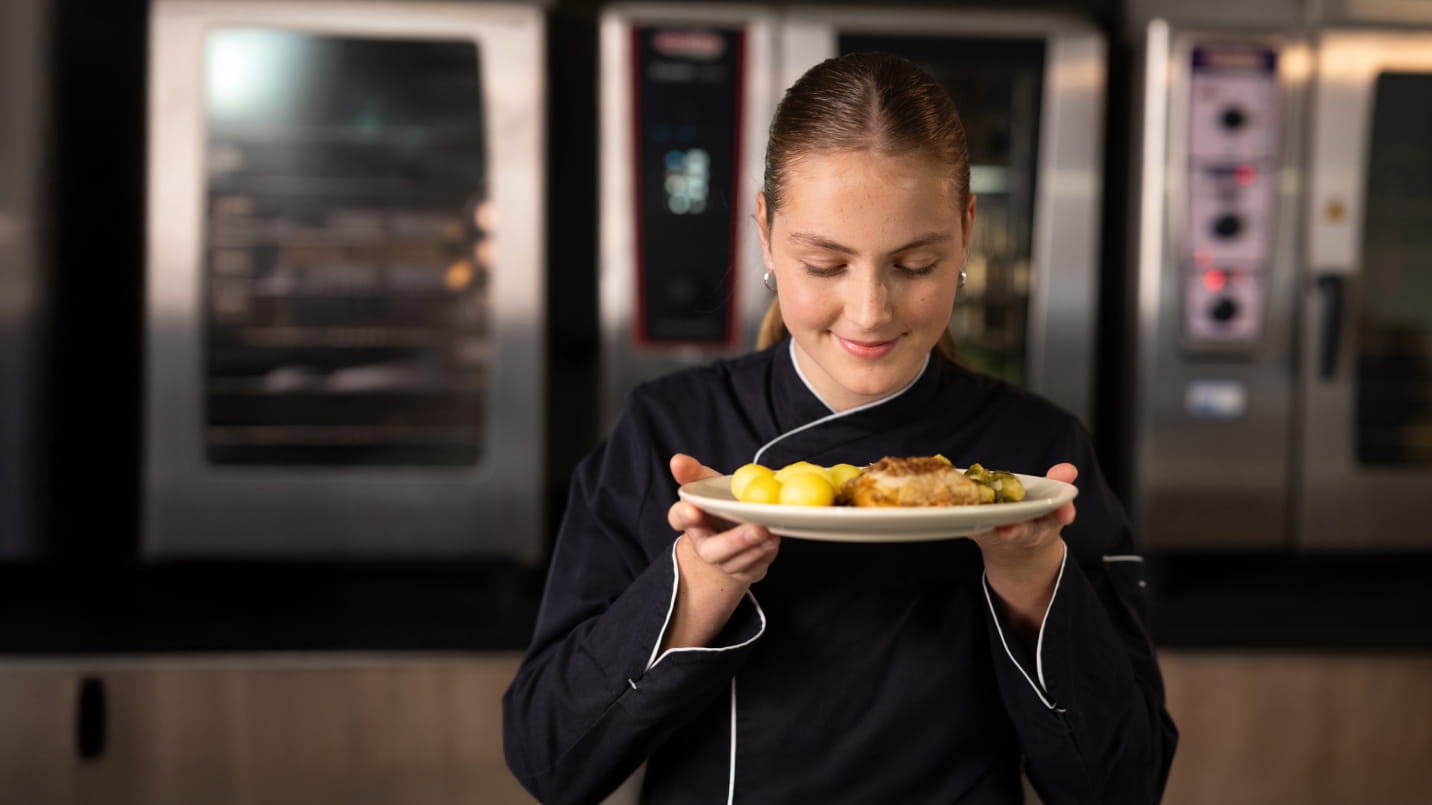 Junge Köchin in schwarzer Uniform mit weißem Kragen hält lächelnd einen Teller mit goldgelben Kartoffeln, paniertem Fleisch und Gemüse in den Händen. Im Hintergrund unscharfe Profi-Öfen einer Großküche.