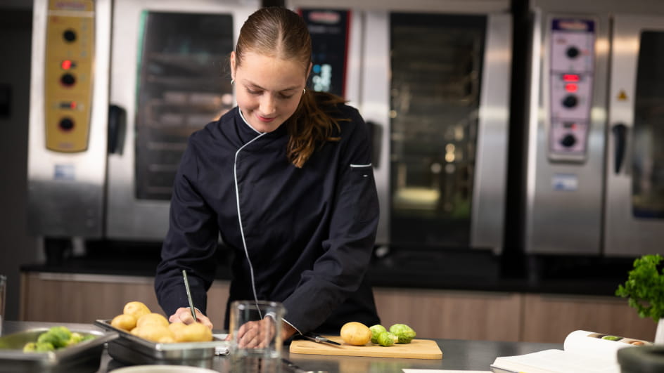 Junge Köchin in schwarzer Uniform mit weißem Kragen steht in Profiküche, schneidet Kartoffeln auf einem Holzbrett mit Messer. Vor ihr Schale mit Rosenkohl, Glas und offenes Rezeptbuch. Im Hintergrund Edelstahlöfen.