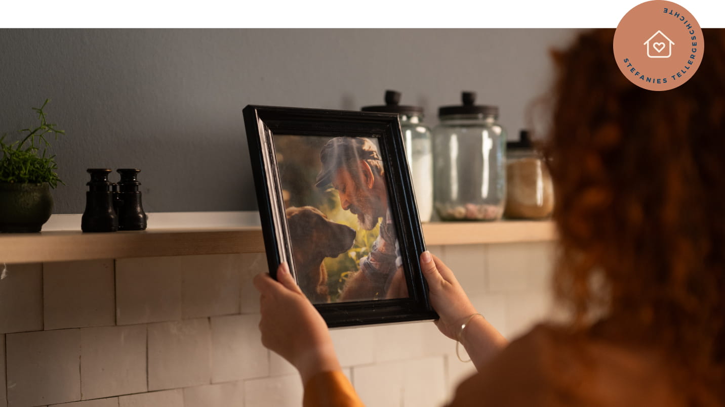 Frau mit lockigen roten Haaren hält von hinten gesehen ein gerahmtes Foto eines älteren Mannes mit Hund. Im Hintergrund Küchenregal mit Gewürzgläsern, Fernglas und kleiner Pflanze. Rechts oben roséfarbenes rundes Badge mit Icon „Haus mit Herz“ und Text „STEFANIES TELLERGESCHICHTE“.