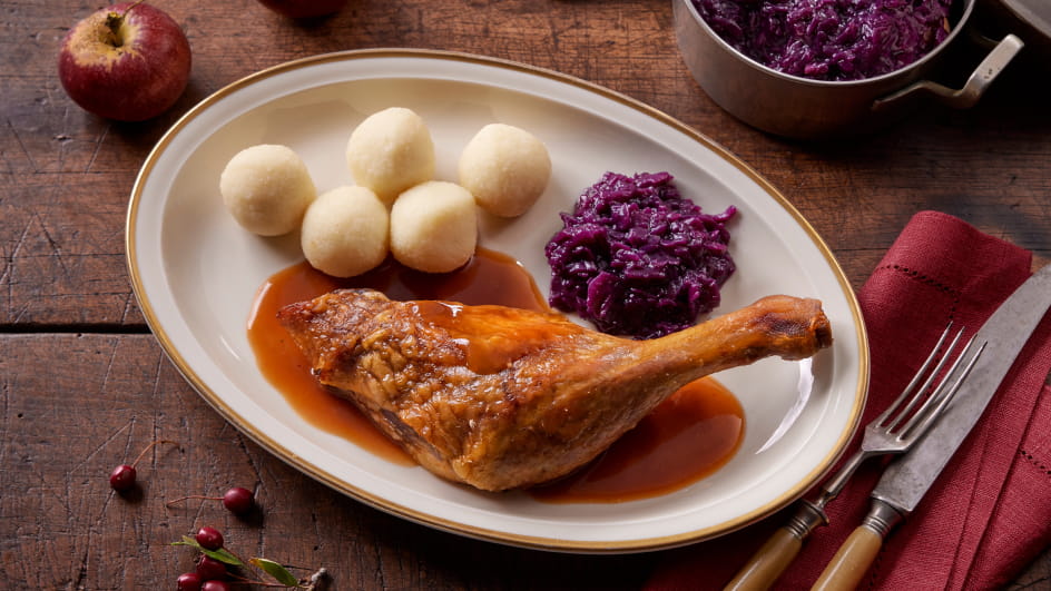 Weißer ovaler Teller mit goldbrauner Entenkeule in Bratensoße, fünf Kartoffelklößen und Rotkohl. Rechts Besteck auf roter Serviette, oben kleine Schale mit mehr Rotkohl, links halber Apfel und Preiselbeeren. Holzuntergrund.