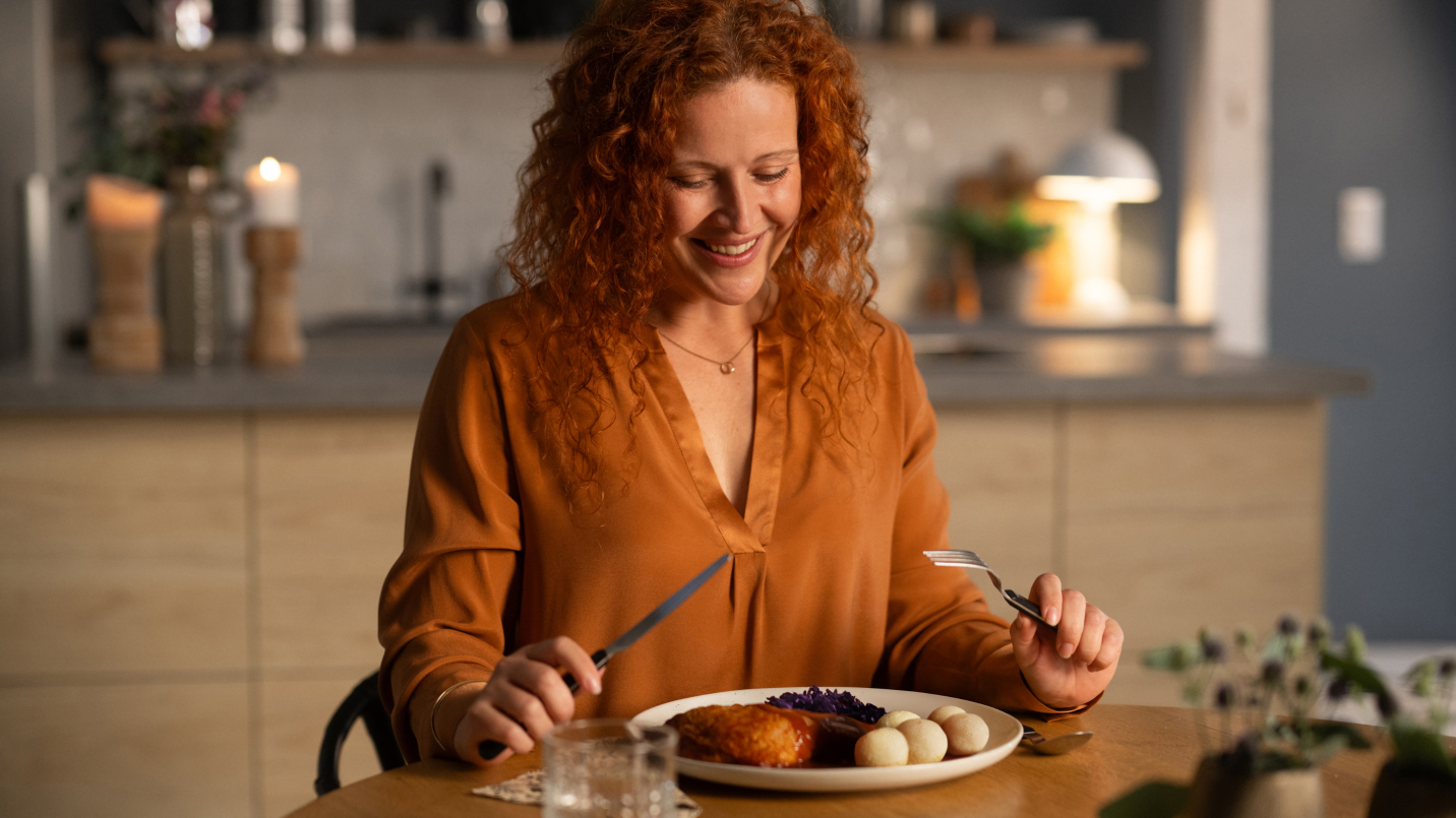 Frau mit lockigen roten Haaren in brauner Bluse sitzt lächelnd am Holztisch, schneidet mit Messer und Gabel ein Gericht aus paniertem Fleisch, Rotkohl und Klößen. Vor ihr Glas Wasser und Löffel. Im Hintergrund moderne Küche mit Kerzen, Blumen und warmer Beleuchtung.