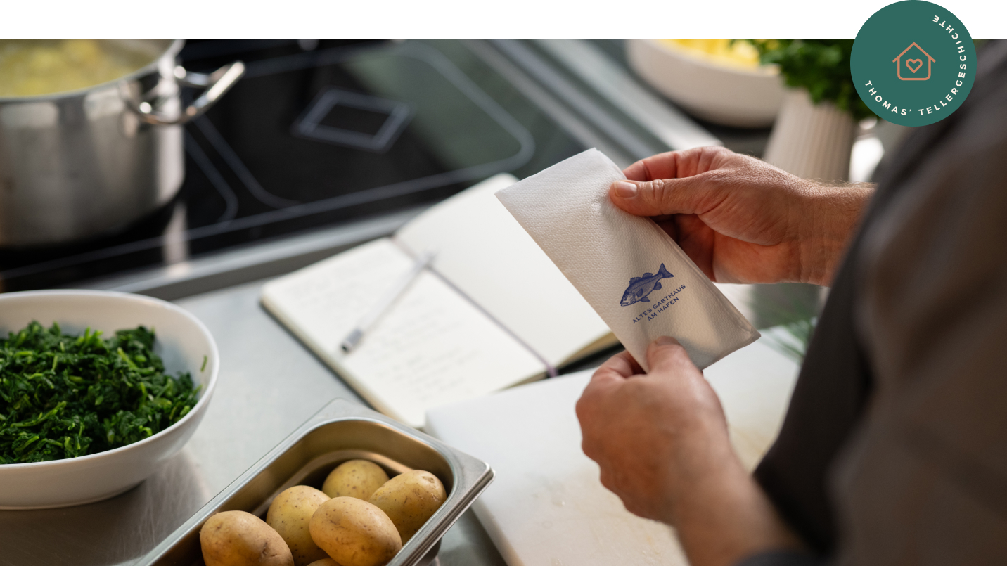 Hände eines Kochs halten eine weiße Serviette mit blauem Fisch-Logo und Text „ALTES GASTHAUS AM HAFEN“. Vor ihm offenes Notizbuch, Schale mit Spinat, Metallbehälter mit Kartoffeln und Schneidebrett. Im Hintergrund Herd mit Topf und Schüssel mit Petersilie. Rechts oben grünes rundes Badge mit Icon „Haus mit Herz“ und Text „THOMAS’ TELLERGESCHICHTE“.