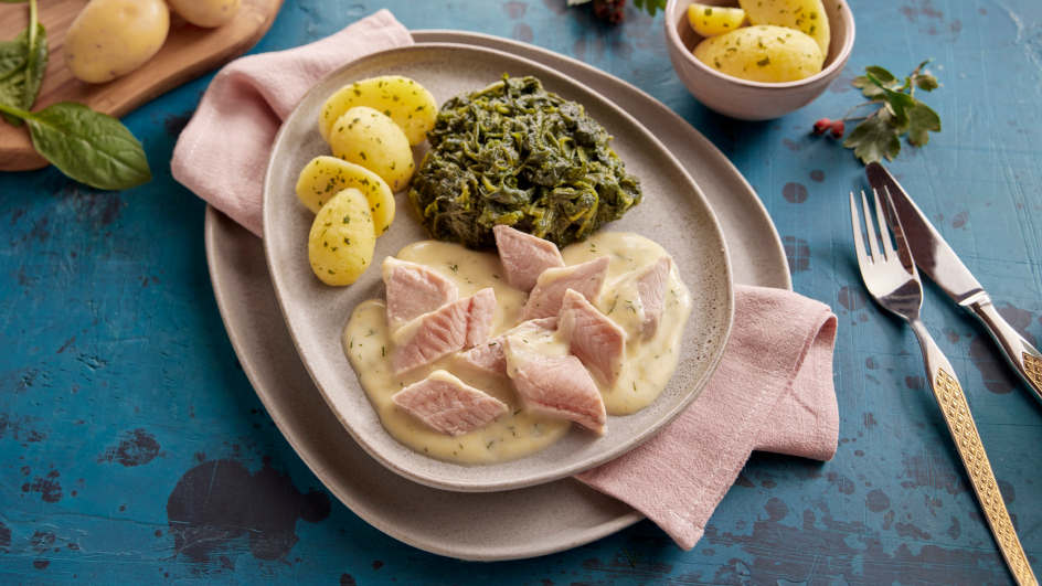 Grauer Teller auf rosa Serviette mit Wildlachs in Weißwein-Sahnesoße, Spinat und Petersilienkartoffeln. Rechts Besteck, oben Schale mit weiteren Kartoffeln. Blauer Tisch mit Spinatblatt und Hagebuttenzweig.