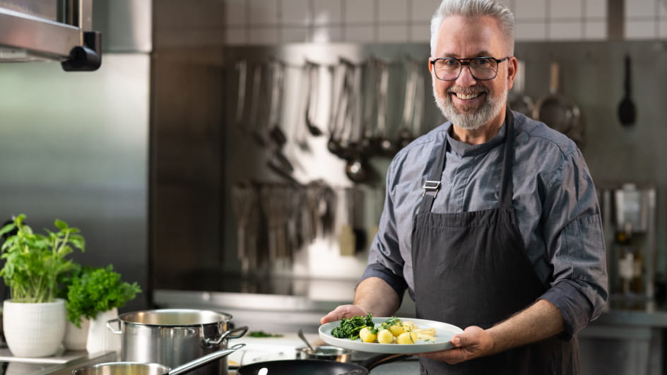 Älterer Koch mit grauem Haar, Bart und Brille in grauer Kochjacke hält lächelnd einen Teller mit Petersilienkartoffeln und Spinat. Im Hintergrund Profiküche mit Edelstahlwänden, hängenden Utensilien und Basilikumtöpfen.